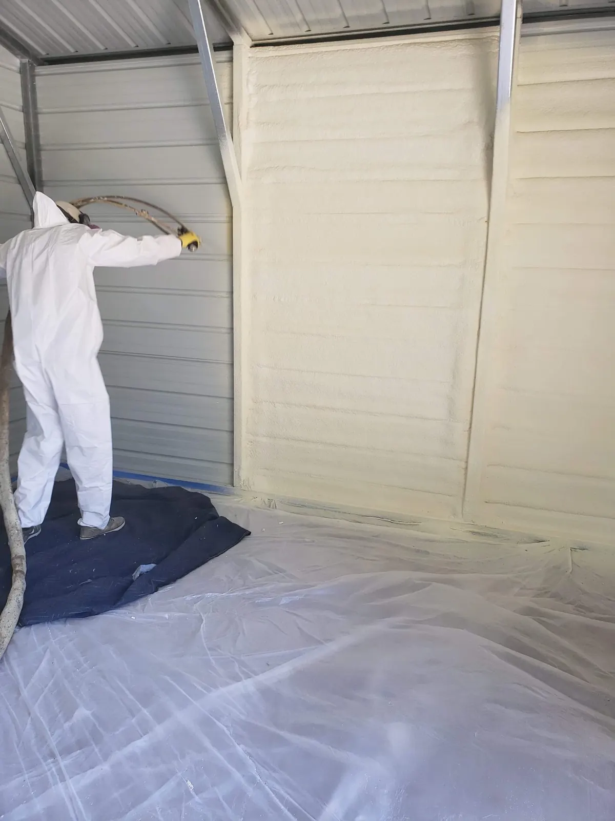 Installer in protective equipment spraying foam onto the interior wall of a metal building, with the floor sheeted for protection
